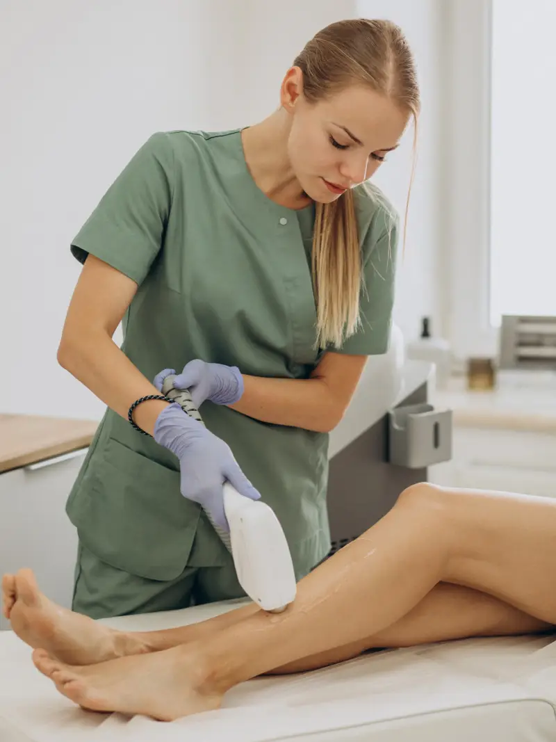 A technician in green scrubs uses a laser device on a client's leg during a hair removal treatment in a bright clinic setting.