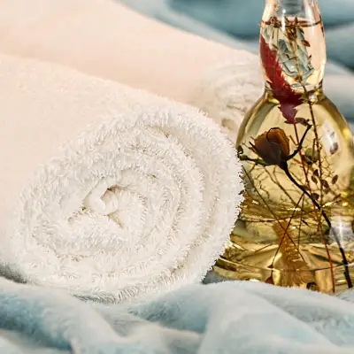 Rolled white towels beside a clear glass bottle containing preserved flowers and a golden liquid, set on a soft, blue surface.